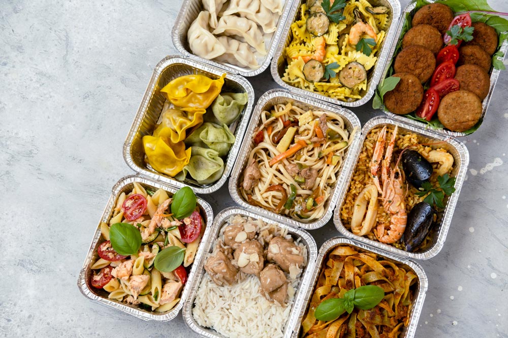 Nio aluminiumlunchlådor med olika maträtter som dumplings, nudlar, ris, pasta och falafel