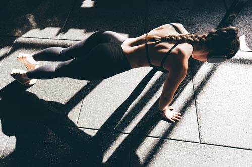 Atletisk kvinna i mörka träningskläder och sport-bh håller en plankposition på ett gymnasiegolv