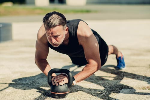 En medelålders muskulös man utför en plankställning med armhävning på en kettlebell utomhus i strålande solsken